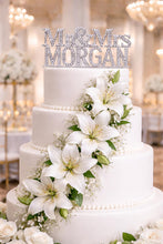 Load image into Gallery viewer, May include: A three-tiered white wedding cake adorned with white lilies and baby's breath. A silver cake topper reads "Mr. & Mrs. MORGAN". The cake is a classic design, ideal for a wedding celebration.