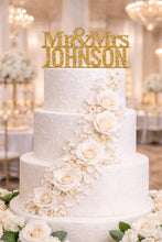 Load image into Gallery viewer, May include: A three-tiered white wedding cake with floral decorations and a gold glitter "Mr. & Mrs. Johnson" cake topper. The cake is decorated with white roses and delicate floral accents, ideal for a wedding celebration.