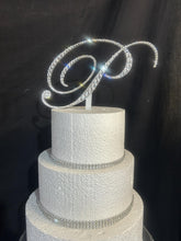 Load image into Gallery viewer, May include: A three-tiered white cake with silver rhinestone bands. A sparkling, cursive letter "P" cake topper, encrusted with rhinestones, sits atop the cake. The cake is set against a dark background.
