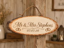 Load image into Gallery viewer, Personalised rustic wood slice wedding sign engraved with “Mr &amp; Mrs Stephens” and wedding date “07.07.26”, finished with joined wedding ring details on each side. The natural bark-edge plaque hangs from rope against a neutral rustic venue background.
